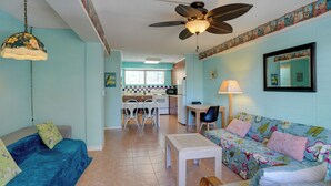Sala de estar com sofá, mesa de centro e ventilador de teto. É visível uma cozinha ao fundo, com mesa e cadeiras.