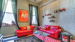 Un salon avec des canapés en cuir rouge, une table basse, un tapis à motifs et une bibliothèque remplie de livres et de jeux de société.