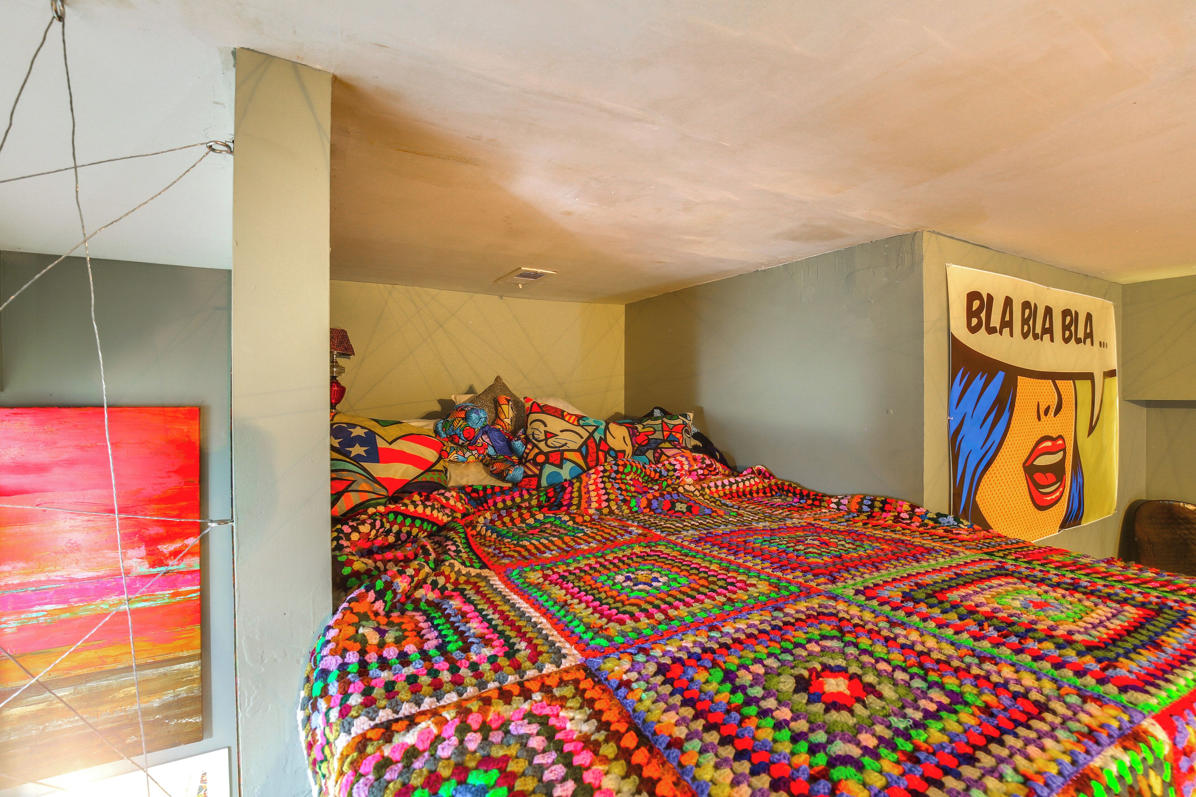 Un dormitorio con una colcha de cama colorida y estampada, un cuadro abstracto grande en la pared y un cartel con texto llamativo.