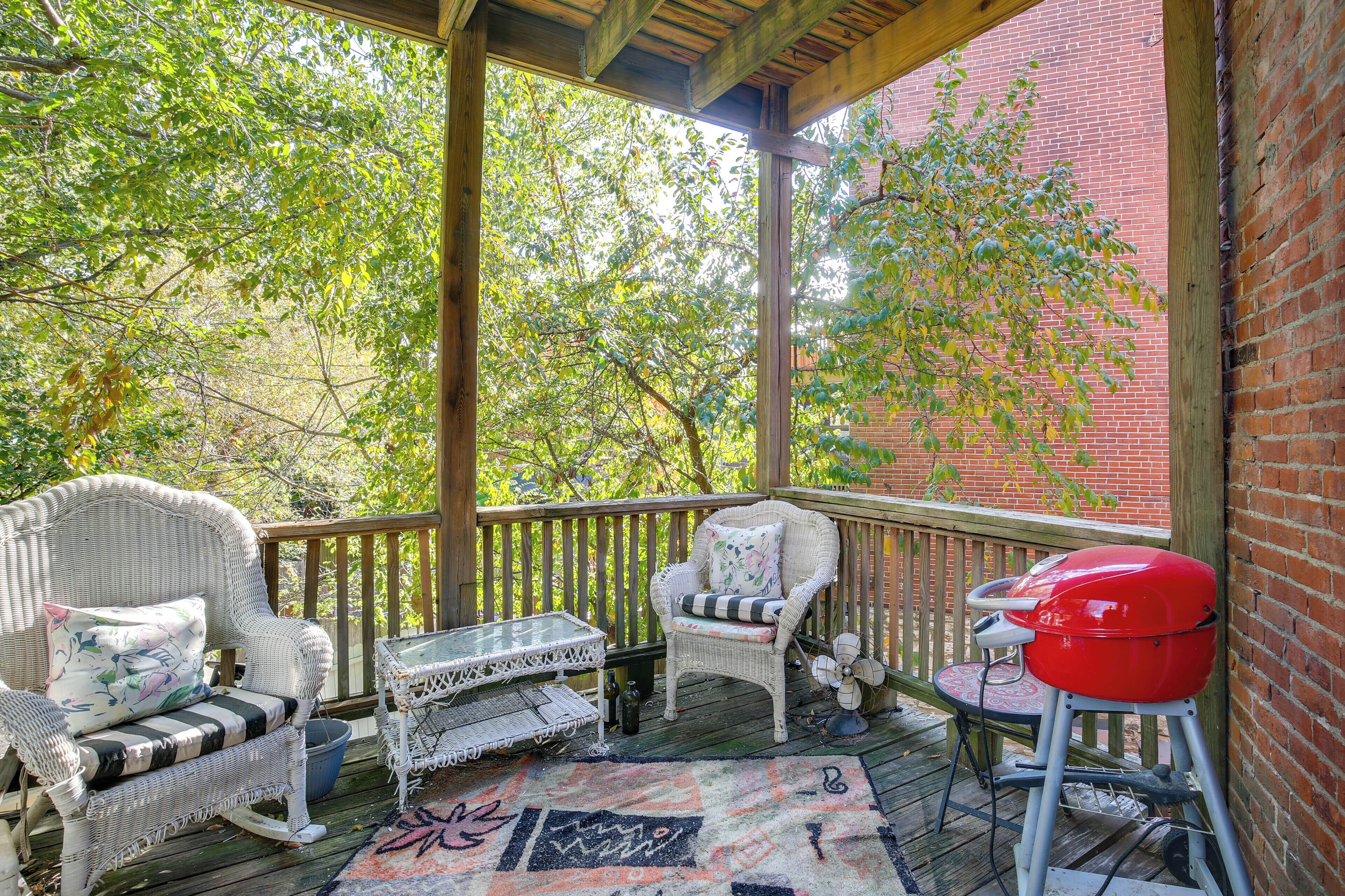 A covered porch with wicker furniture, a red grill, and a view of trees and a brick wall.