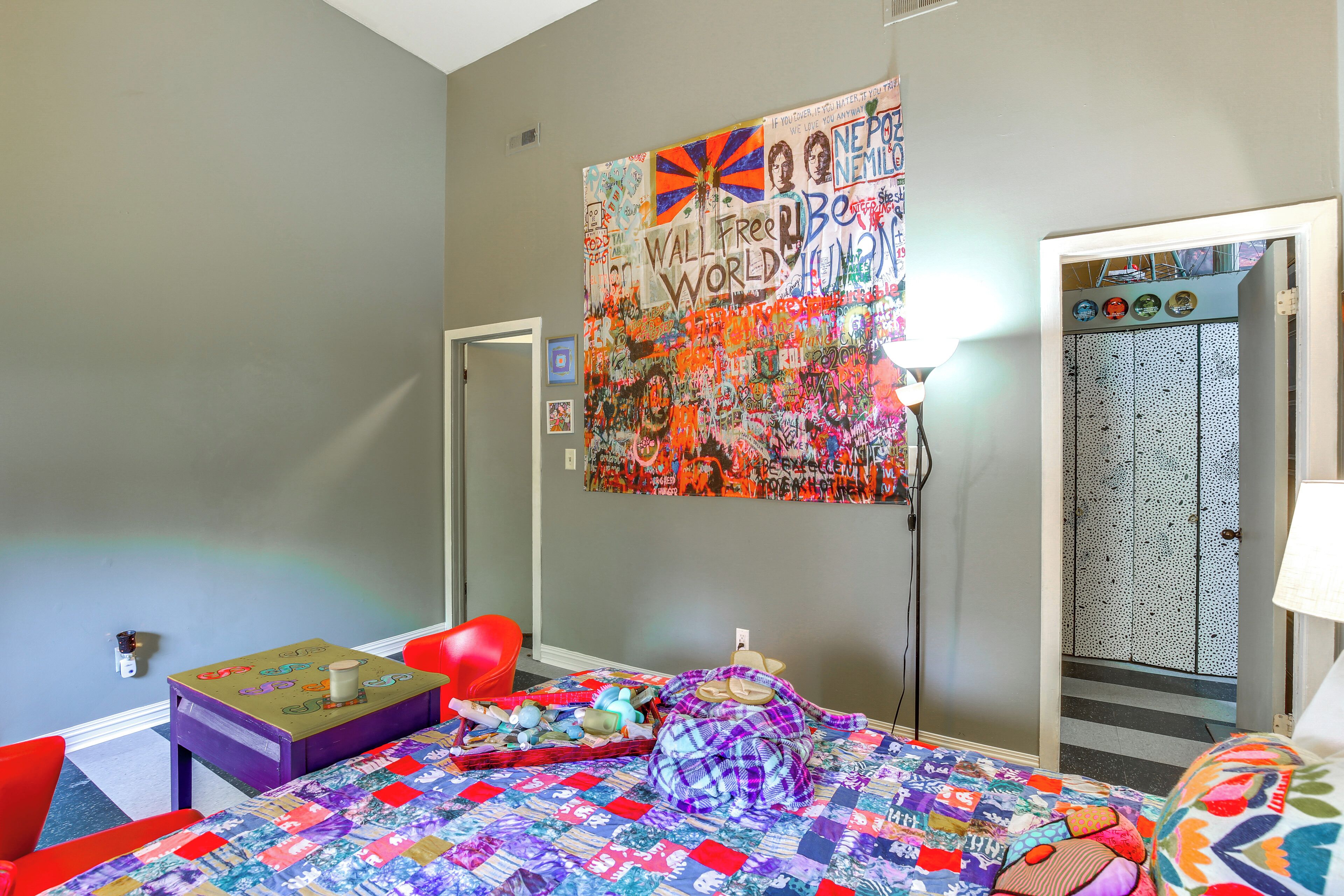 A bedroom with a colorful quilt, a red chair, a small table with a cup, and a door with a patterned panel.