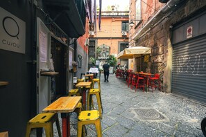 A narrow alley with tables and chairs, a closed shutter on the right, and a building with a sign reading "CUQU". - Song House Apartment, Historic Center of Naples (Napoli)