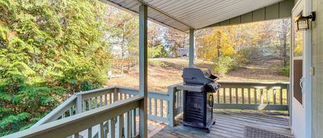 A covered porch with a grill, wooden deck, and a view of a forested area.