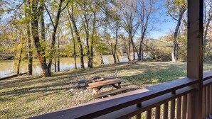 A wooden deck with a picnic table and chairs overlooking a lake, surrounded by trees and a distant building. - Cabin Along Rough River! Views, Fire Pit & Grill (Falls of Rough)