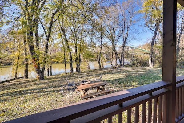 A wooden deck with a picnic table and chairs overlooking a lake, surrounded by trees and a distant building.