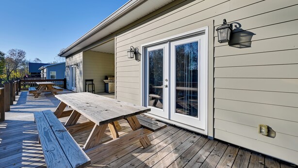 Une terrasse en bois avec des tables de pique-nique, un espace couvert et une maison dont le revêtement extérieur est de couleur claire et qui possède une porte-fenêtre coulissante.