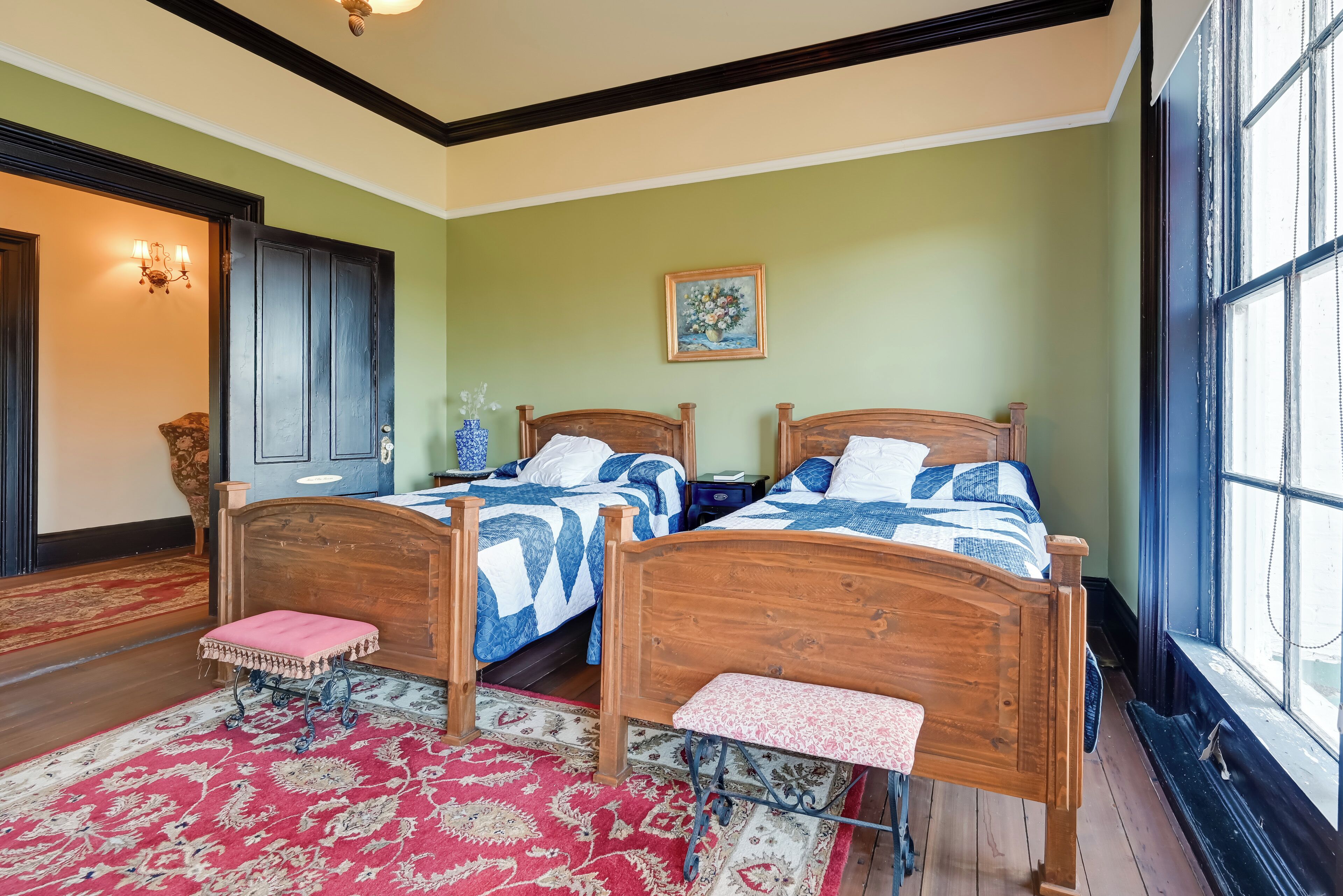 Une pièce avec deux lits en bois, un tapis rouge à motifs, un banc en bois, un lustre et une grande fenêtre.