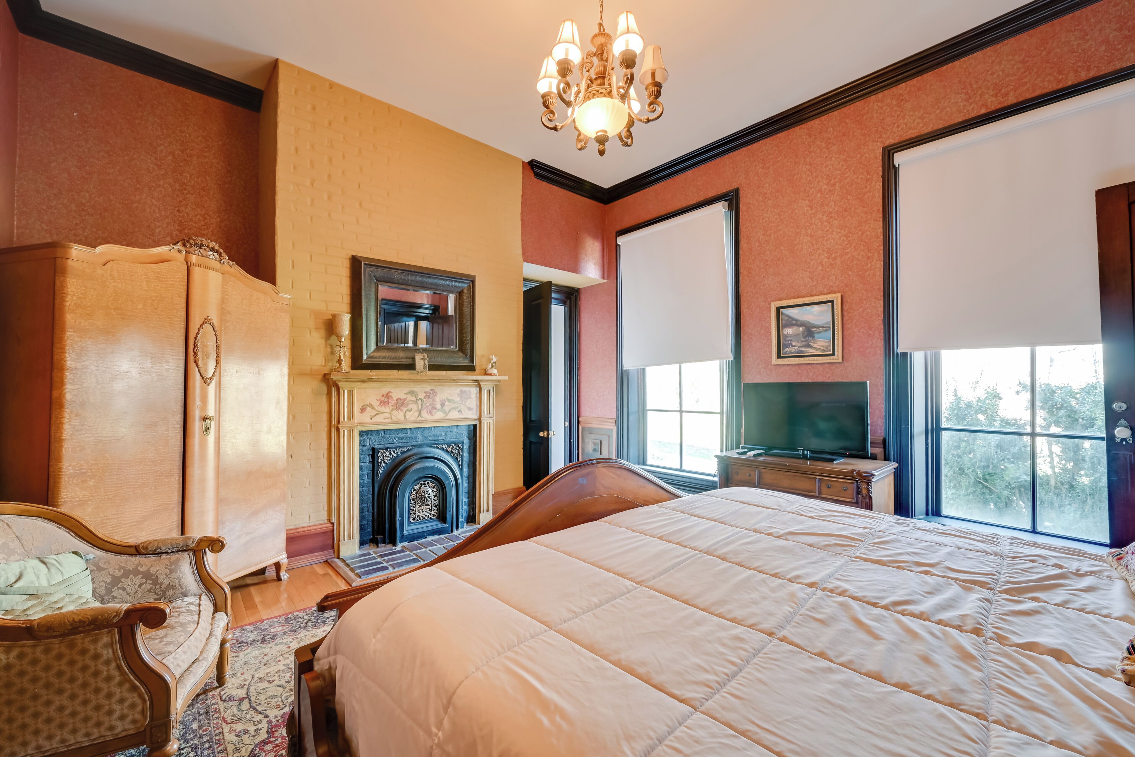 A bedroom with a large bed, a wooden wardrobe, a fireplace, a television, and a window with a view.