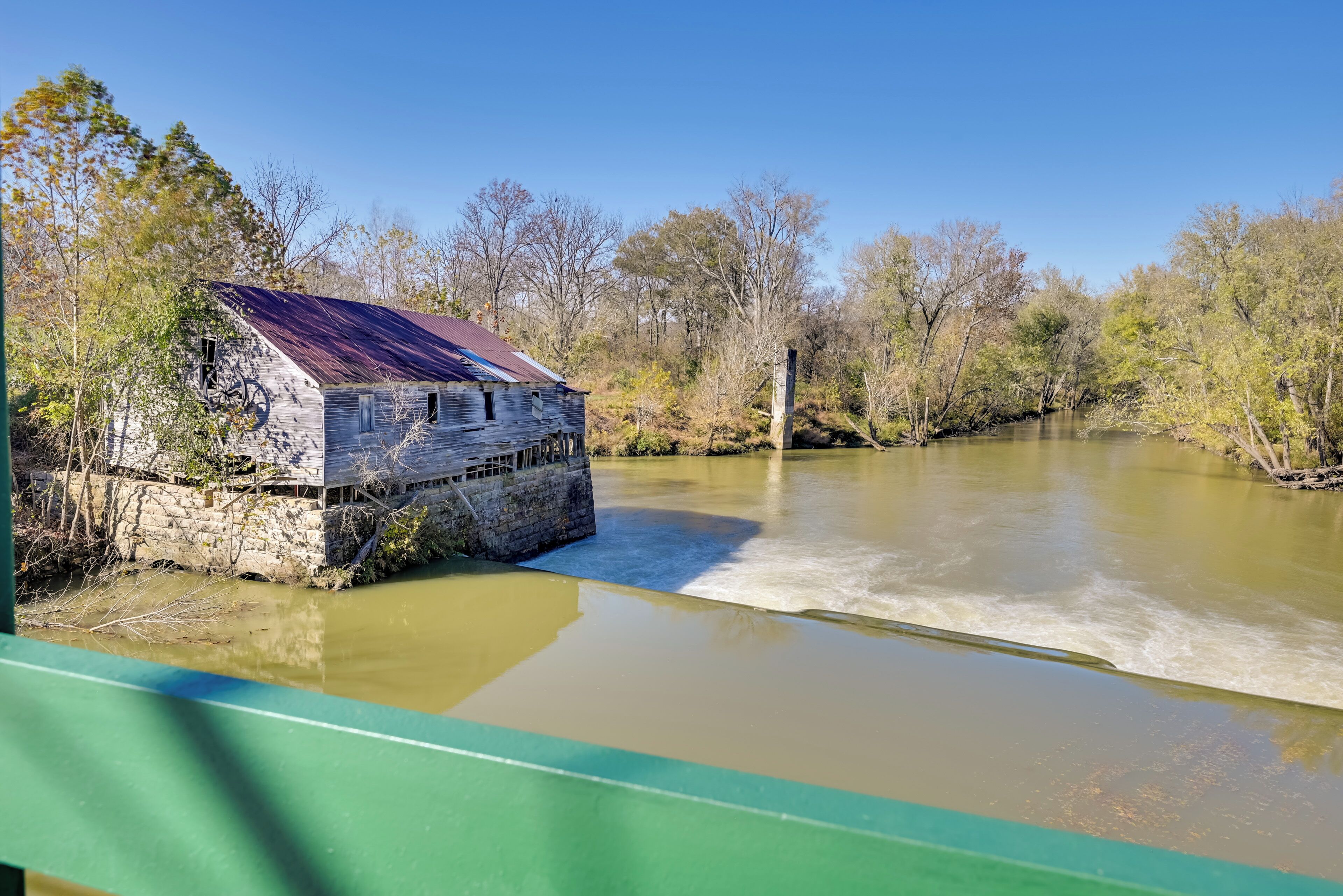 A rustic wooden building by a calm river, surrounded by trees and a clear sky.