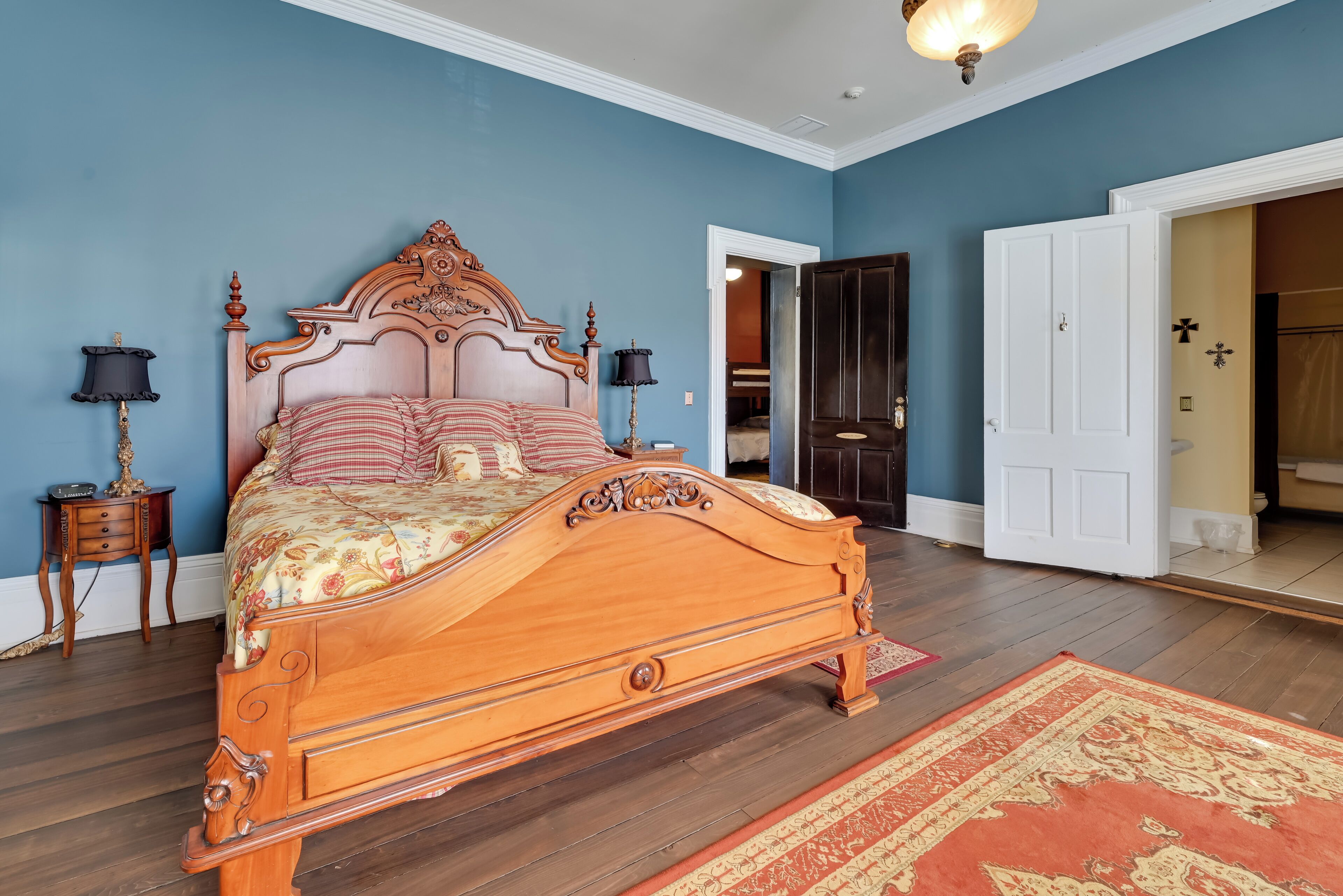A bedroom with a wooden bed, two bedside tables with lamps, a patterned rug, and a door leading to another room.
