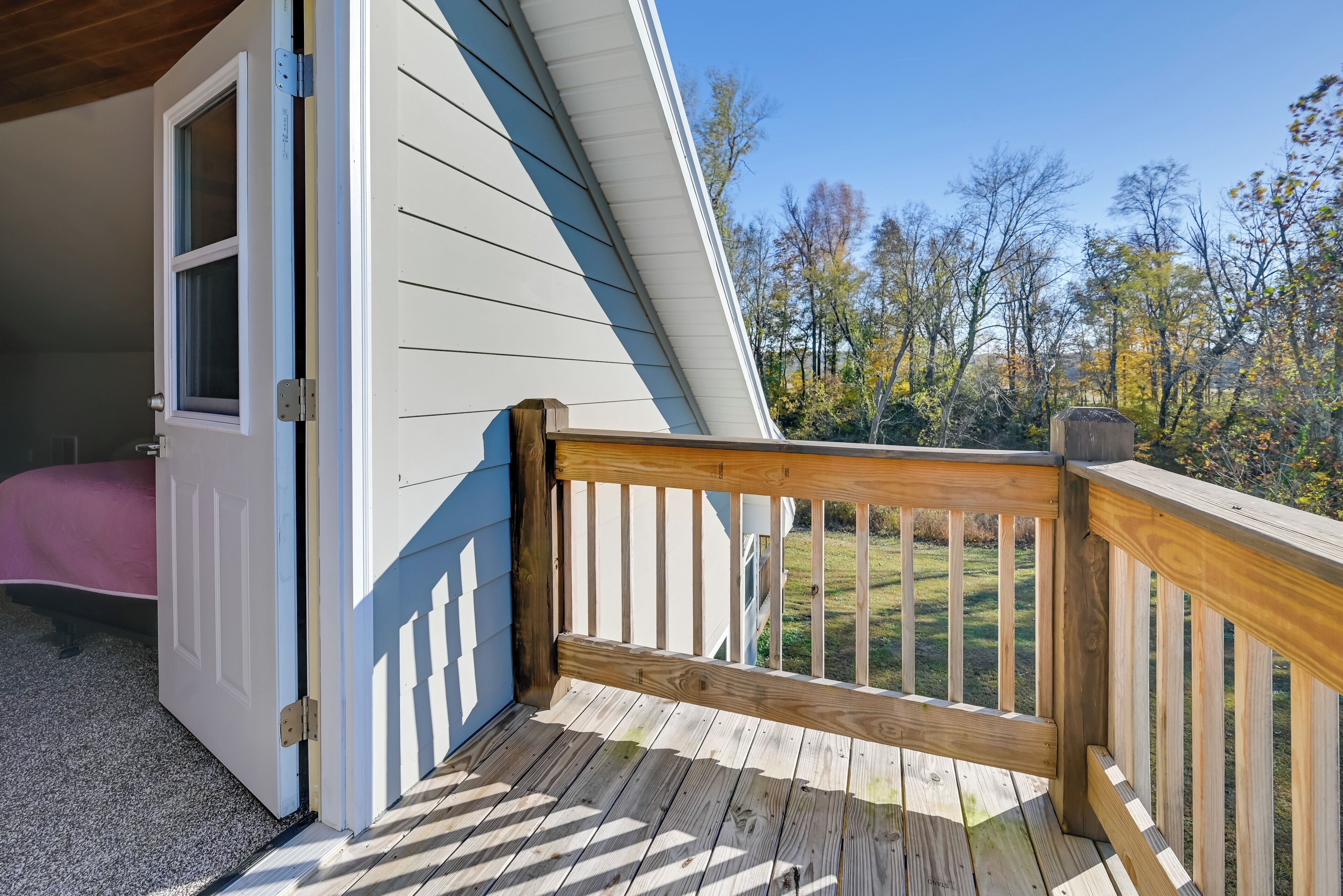 A wooden deck with a white railing, a door leading to a bedroom, and a view of a grassy area with trees.