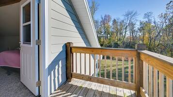 A wooden deck with a white railing, a door leading to a bedroom, and a view of a grassy area with trees.