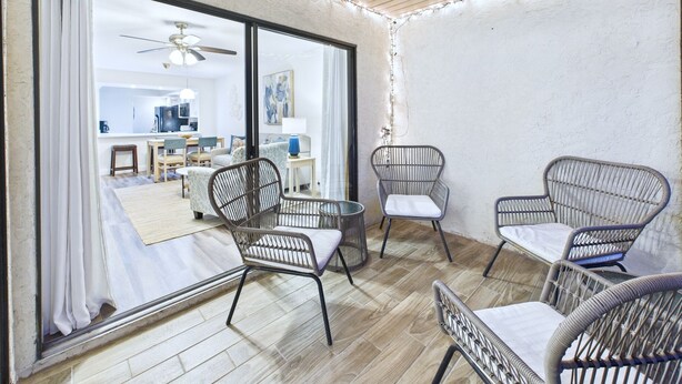 A room with wicker chairs, a ceiling fan, and a view of a kitchen and dining area through large windows.