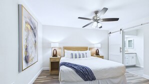 A modern hotel room with a large bed, a ceiling fan, a nightstand with a lamp, and a large mirror.