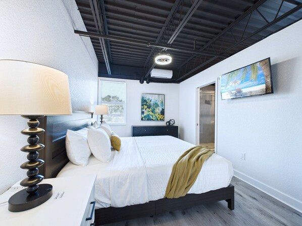 A modern bedroom with a bed, bedside lamps, a painting, and a window. - Experience modern elegance at Mangrove Lofts—steps from Port Canaveral and minutes to Cocoa Beach. Enjoy sleek design, premium comfort, onsite cruise parking avail, easy access to beaches, and rocket launch views from inside unit! (Cape Canaveral)