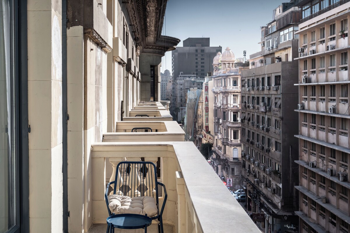 Un balcón estrecho con una silla y cojines, con vistas a un paisaje urbano con varios edificios y balcones.