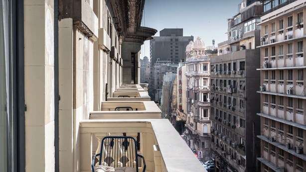 A narrow balcony with a chair and cushions, overlooking a cityscape with multiple buildings and balconies. - Brassbell grey apartments Bidair House (Cairo)
