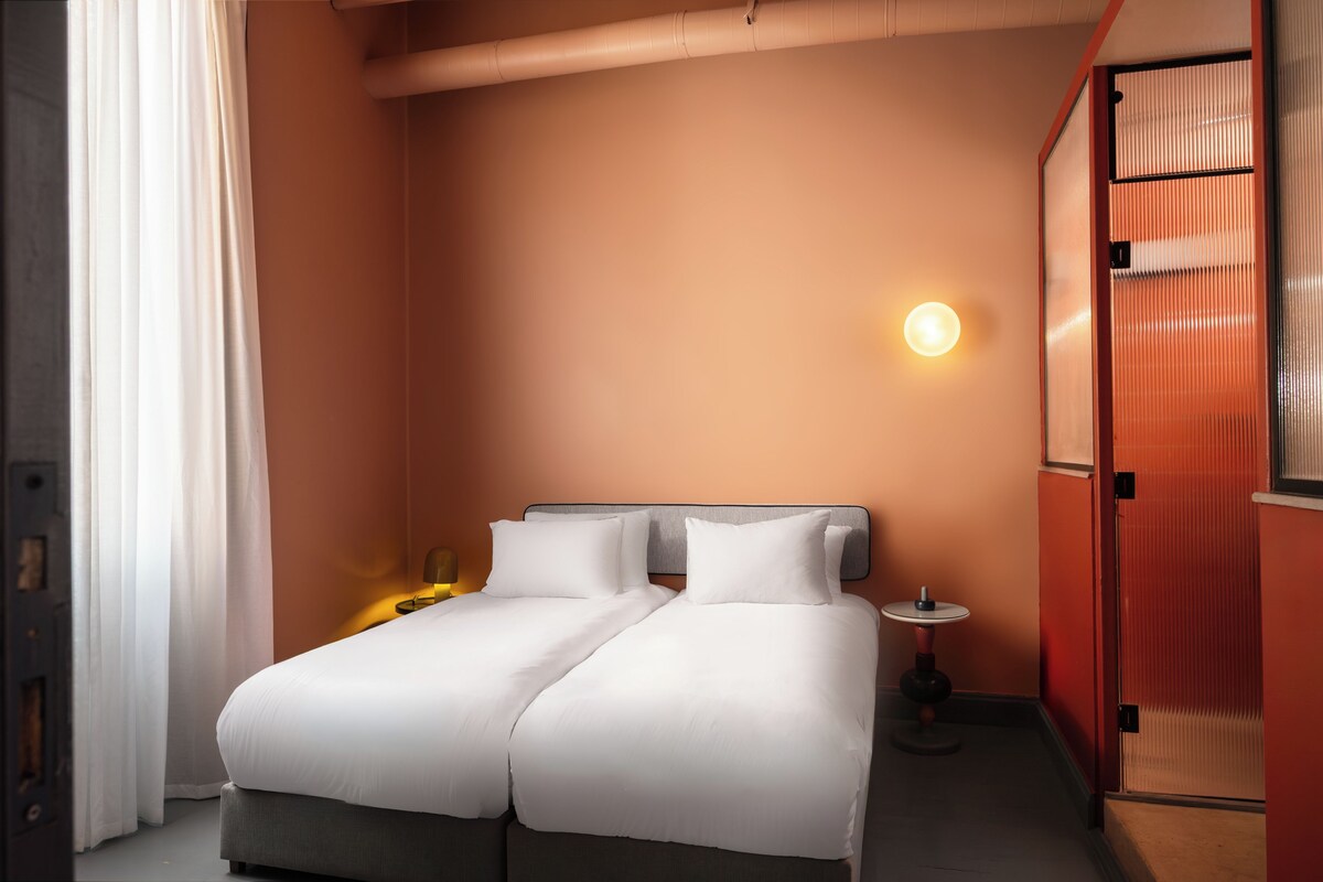 Una cama doble con ropa de cama blanca, una mesita de noche con lámpara y un aplique en la pared.