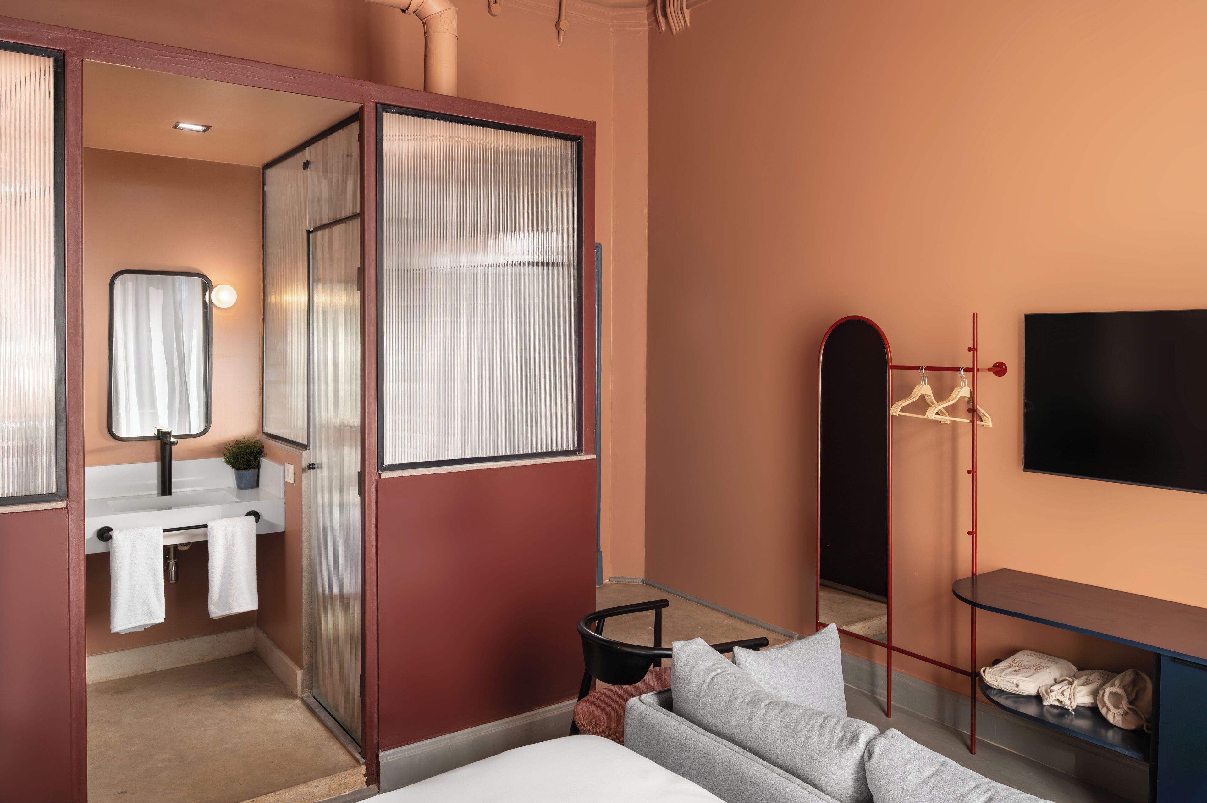 Ett modernt hotellrum med en glasvägg som avskiljer badrummet, en platt-TV som är monterad på väggen, en liten sittgrupp med en soffa och ett bord samt en klädställning med galgar.