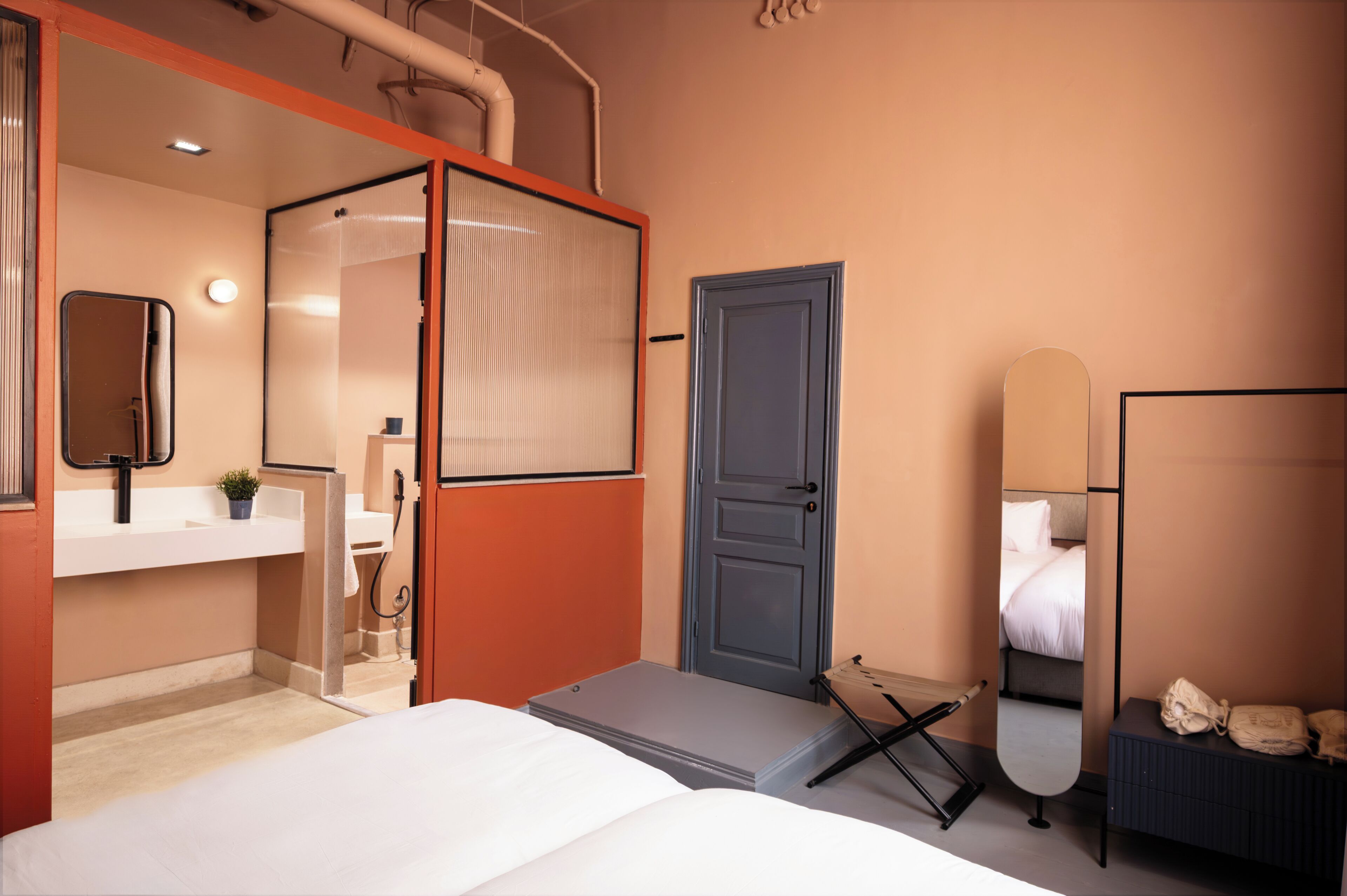 Une chambre d’hôtel de taille réduite, comprenant un lit, un petit bureau, un miroir, une porte et une salle de bain avec un lavabo et un miroir.