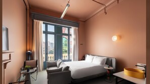 A hotel room with a bed, a sofa, a small table with a plant, a round table with a lamp, and a large window with curtains. - Brassbell grey apartments Bidair House (Cairo)