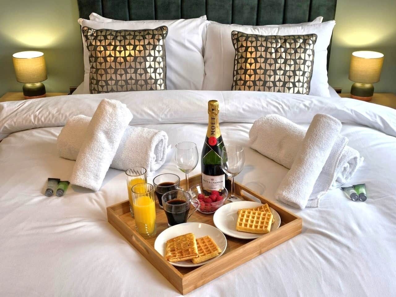 A neatly made bed with white linens, a bottle of champagne, waffles, and juice on a tray.