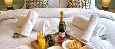 A neatly made bed with white linens, a bottle of champagne, waffles, and juice on a tray.