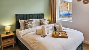 A neatly made bed with pillows, a bedside table with a lamp, a tray with breakfast items, and a window with a view of buildings and a car.