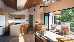 Une cuisine moderne avec des meubles en bois, un îlot central et un coin repas comprenant une table et des chaises. Une terrasse aménagée avec des meubles d’extérieur offre une vue sur la verdure.