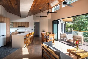 Une cuisine moderne avec des meubles en bois, un îlot central et un coin repas comprenant une table et des chaises. Une terrasse aménagée avec des meubles d’extérieur offre une vue sur la verdure.