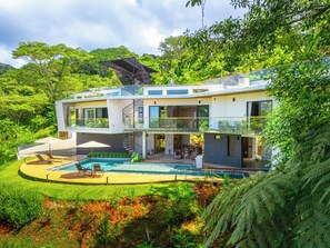 Une maison moderne à deux étages avec une piscine, entourée d’une végétation luxuriante.