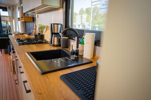 A modern kitchen with a gas stove, sink, coffee maker, and a window with a view of the outside. - Floatinghouse Emsel WOMA D13 – Modernes Hausboot für bis zu 6 Personen am Röblinsee (Fürstenberg)