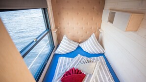 Una habitación con cama, almohadas a rayas, una manta roja y ventana con vista al agua.