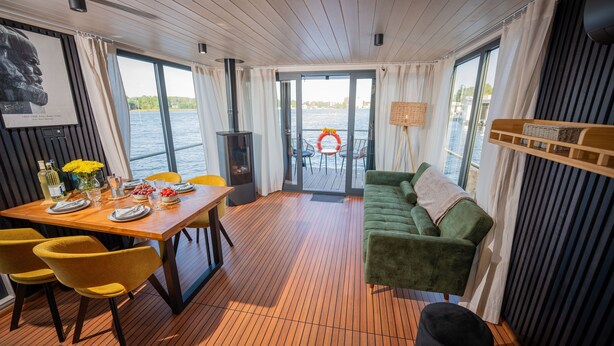 Comedor con una mesa de madera, sillas amarillas, un sofá verde y vistas al agua a través de amplios ventanales.