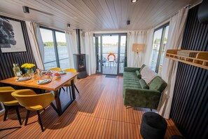 Een eetruimte met een houten tafel, gele stoelen, een groene bank en uitzicht op het water door grote ramen.