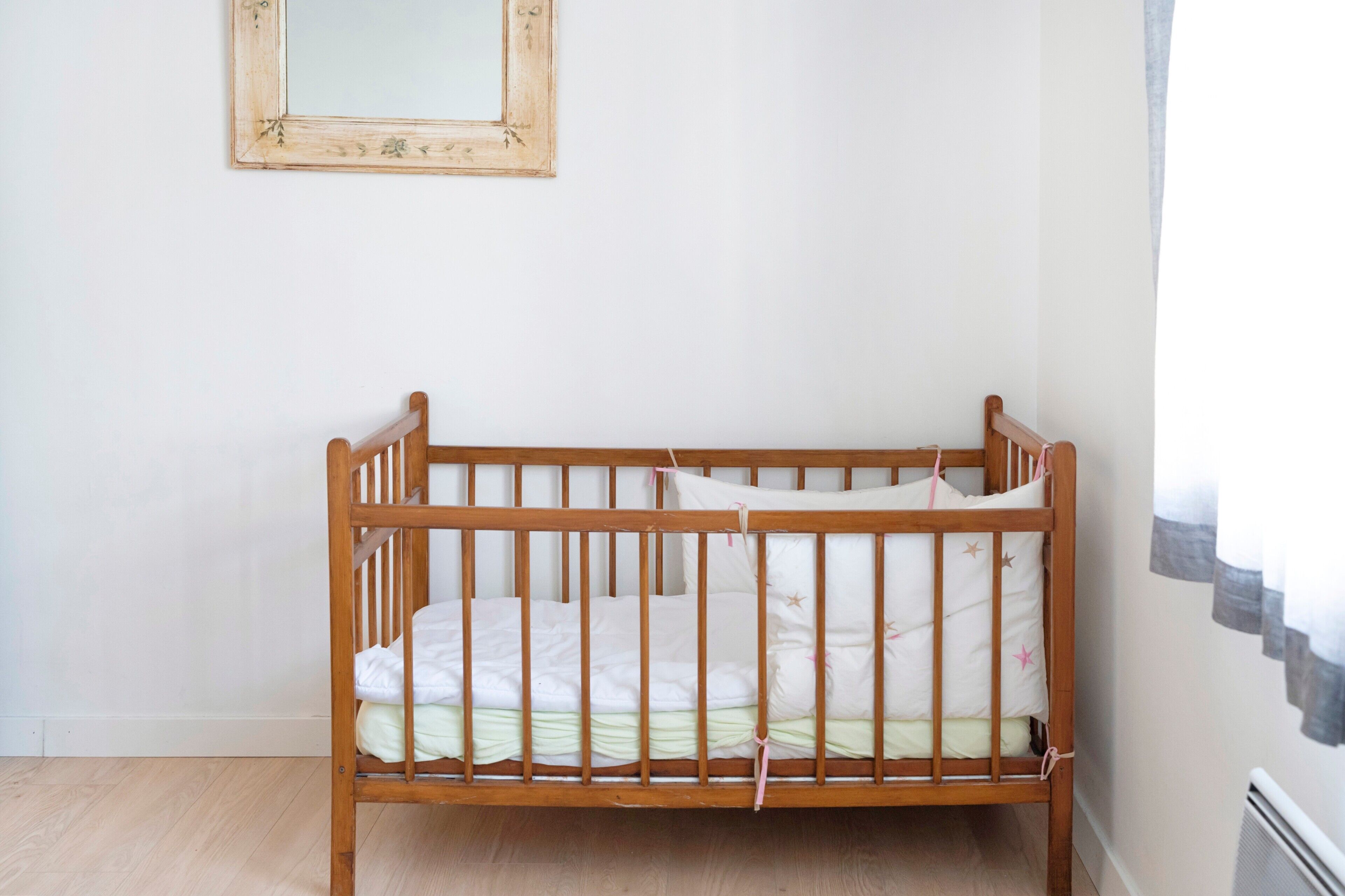 Una cuna de madera con colchón y almohadas, una pared lisa y una ventana con cortinas.