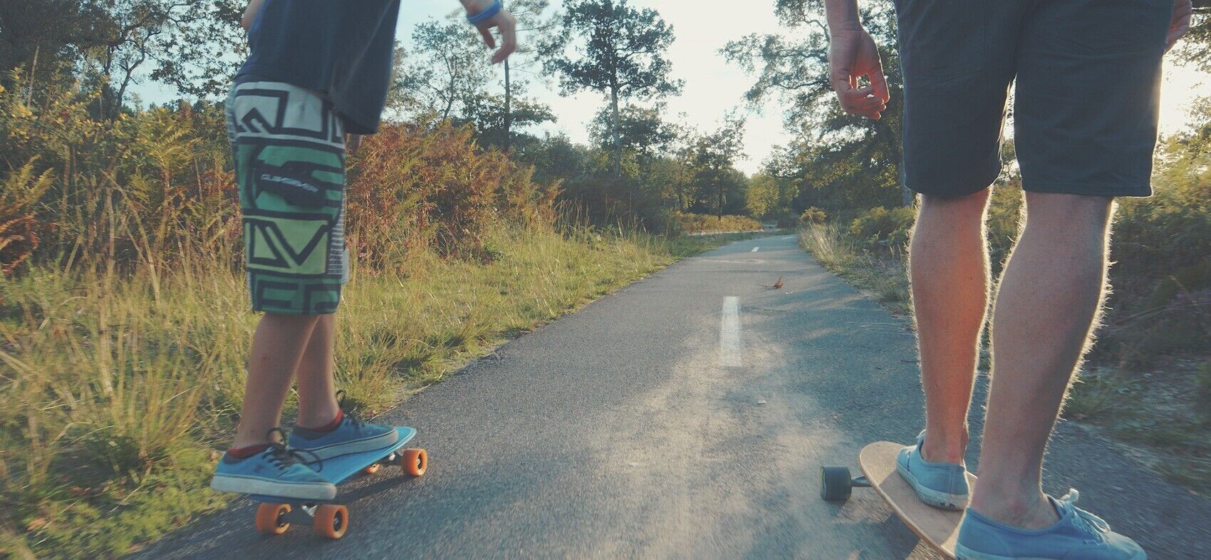 Twee personen skateboarden op een verharde weg met begroeiing aan beide zijden.