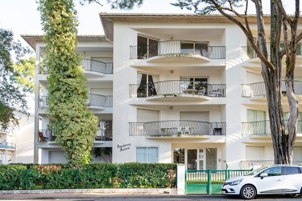 Un immeuble résidentiel à plusieurs étages, doté de balcons, d’une clôture verte et d’une voiture blanche garée devant.