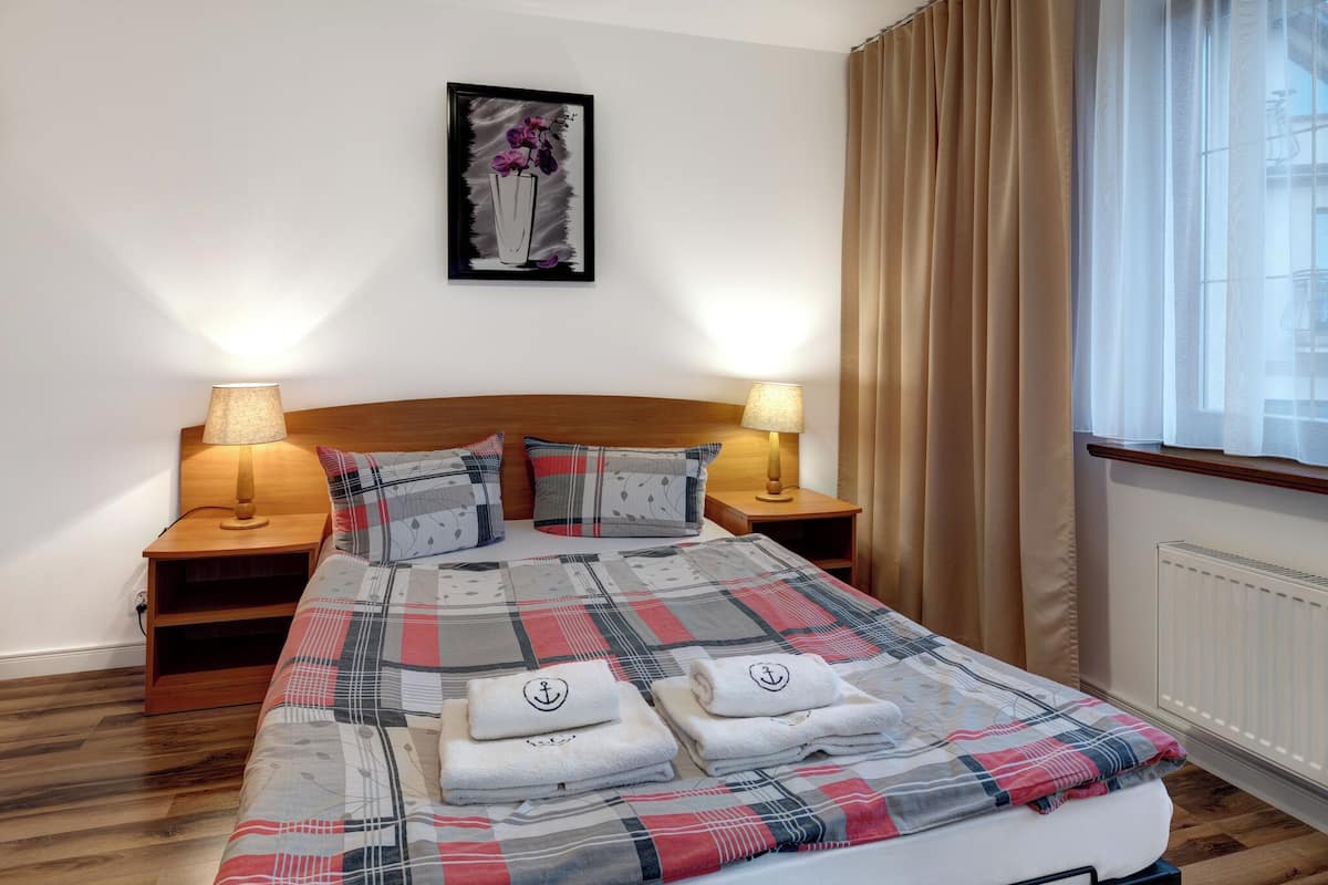 Una cama bien hecha con una colcha escocesa, dos lámparas de noche, una obra de arte enmarcada en la pared y una ventana con cortinas.