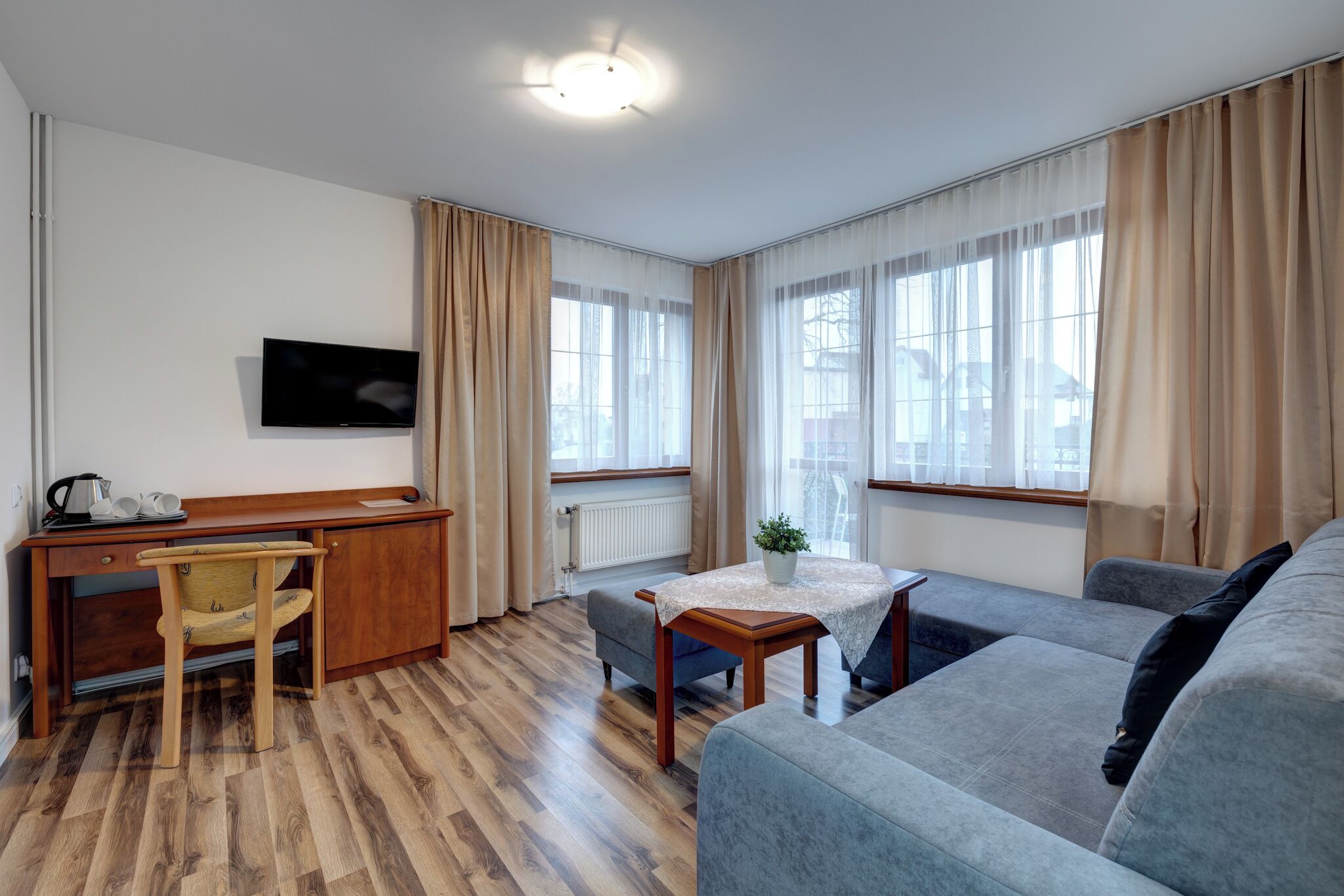 Ett modernt vardagsrum med ett skrivbord i trä, en grå soffa, ett litet bord med en växt och en tv som är monterad på väggen.