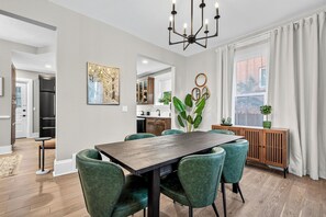 Un espace repas avec une table en bois et des chaises à revêtement vert, un lustre, et une vue sur la cuisine et le salon.