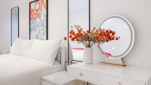 Ett modernt sovrum med en vit säng, ett sängbord med en spegel, en vas med blommor och en inramad affisch med en stadsbild på väggen.