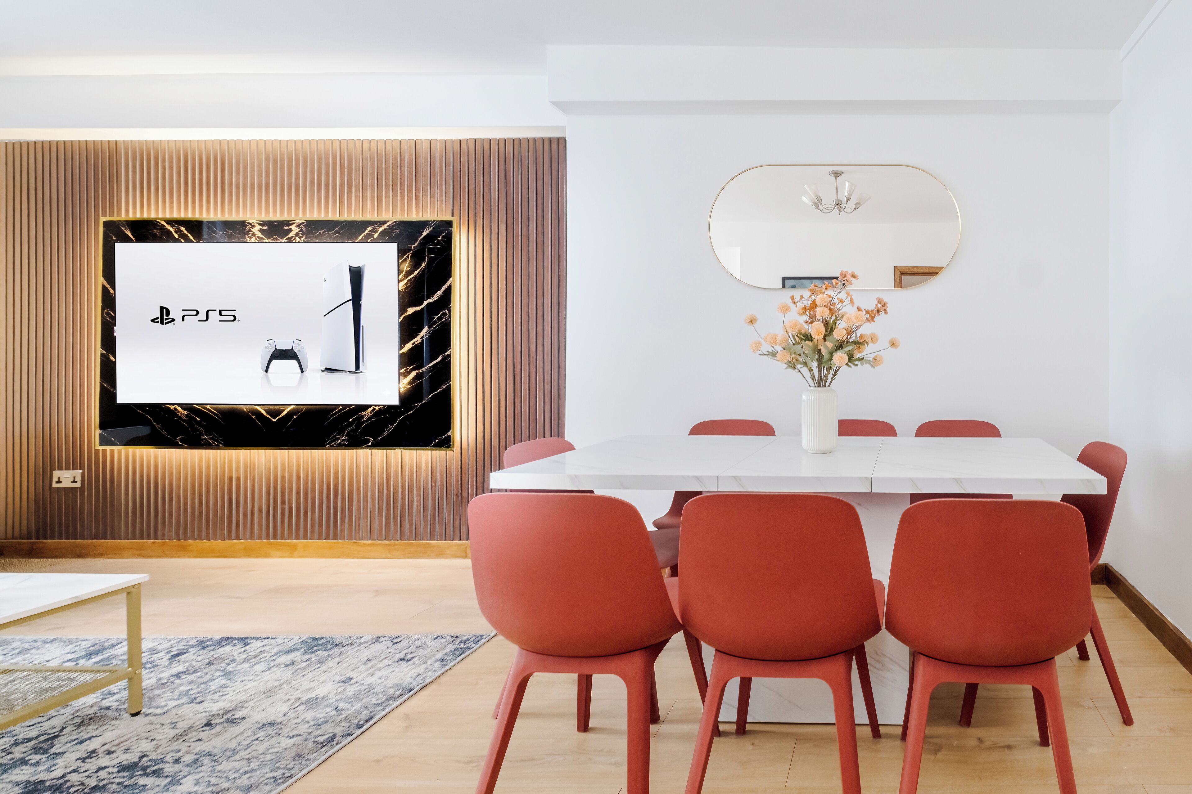 A modern dining area with a white table and red chairs, a large screen displaying the PlayStation logo, and a vase with flowers on the wall.