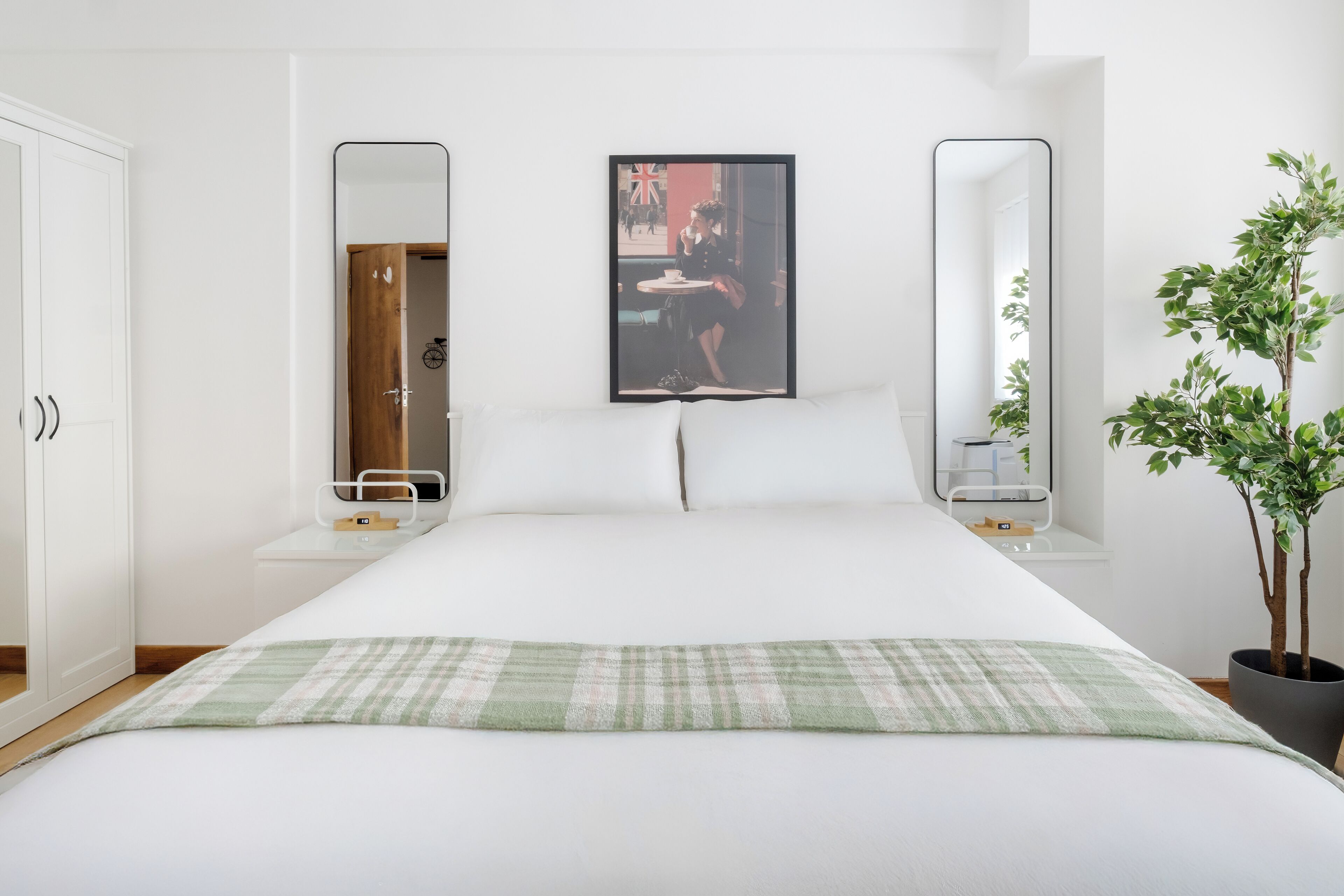 Ein ordentlich bezogenes Bett mit karierter Tagesdecke, zwei Nachttische mit Spiegeln, ein gerahmtes Bild an der Wand und ein Blumentopf.
