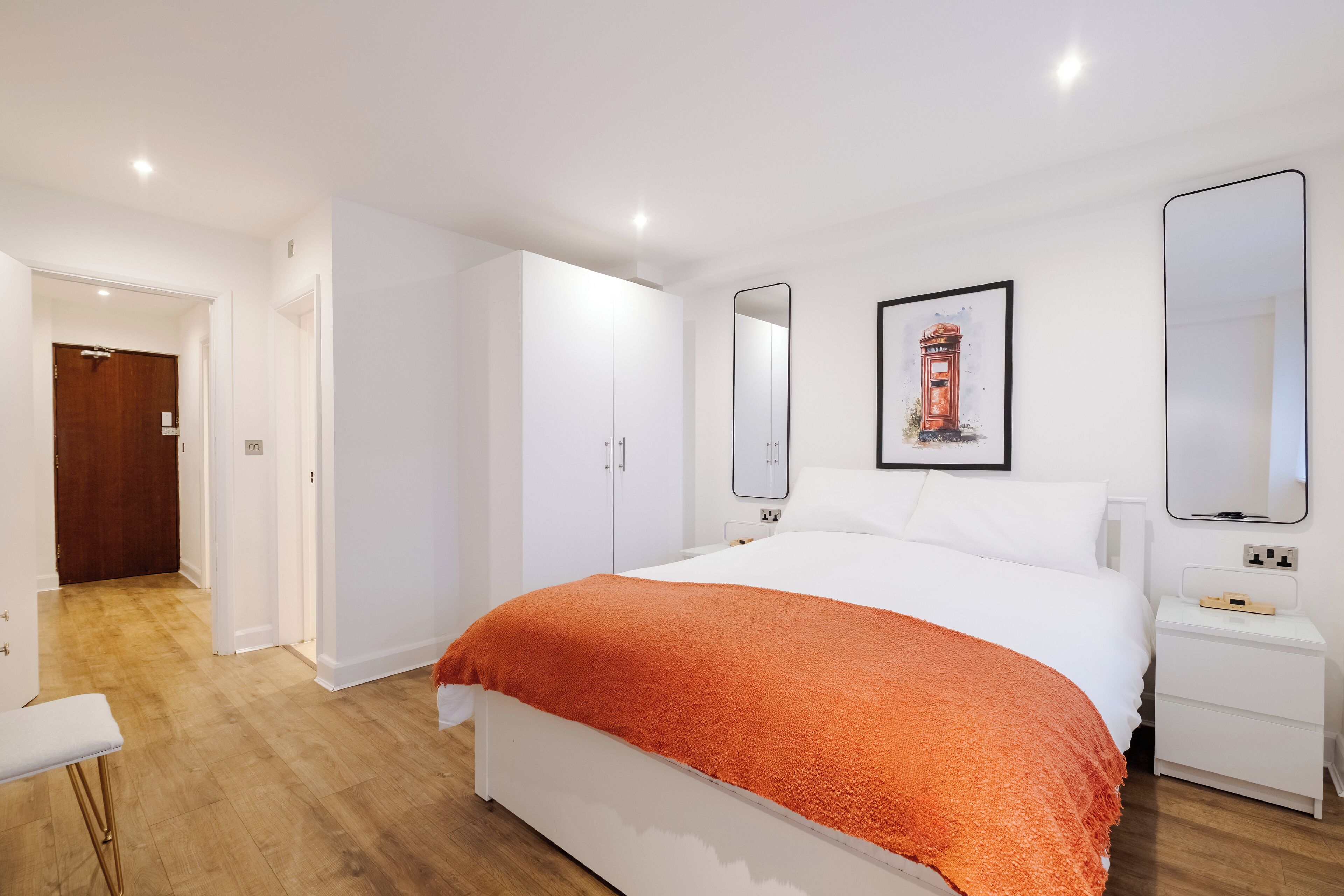 A modern hotel room with a large bed, white walls, and a wooden floor. There is a door leading to another room and a small bedside table with a lamp.