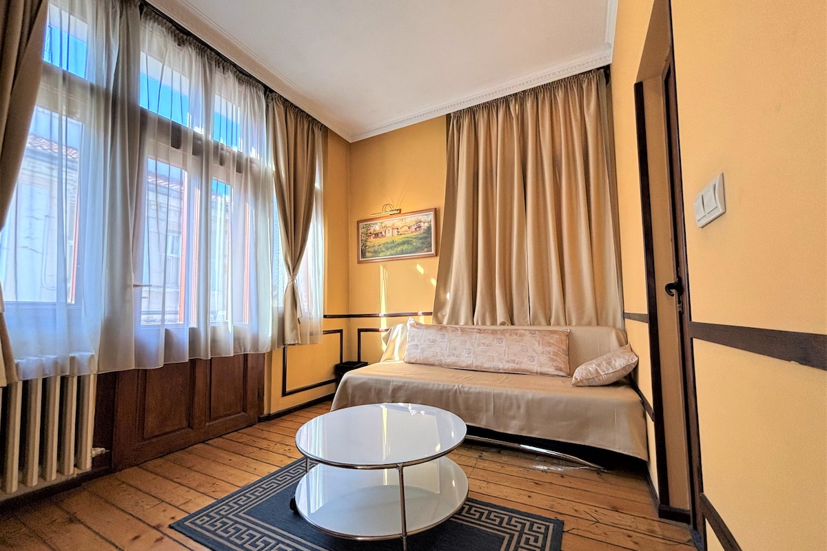 A hotel room with a bed, a small round table, a window with curtains, a painting on the wall, and a wooden floor.