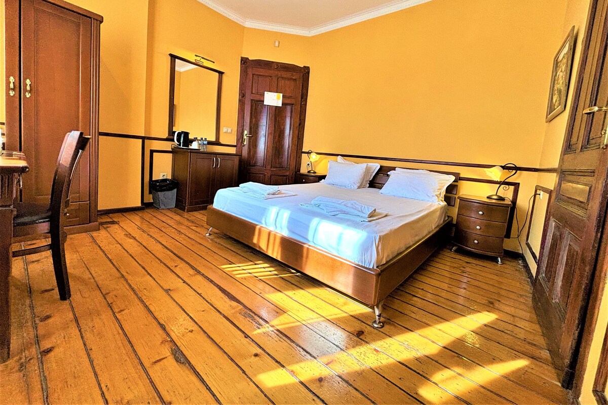 A bedroom with a wooden floor, a bed with white linens, a wooden wardrobe, a desk with a chair, and a mirror on the wall.