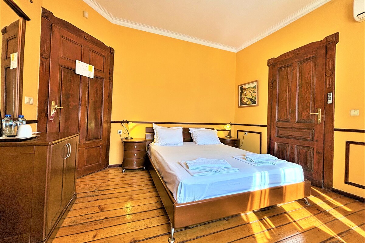 A hotel room with a wooden floor, a bed with white linens, a nightstand with a lamp, and wooden wardrobes.