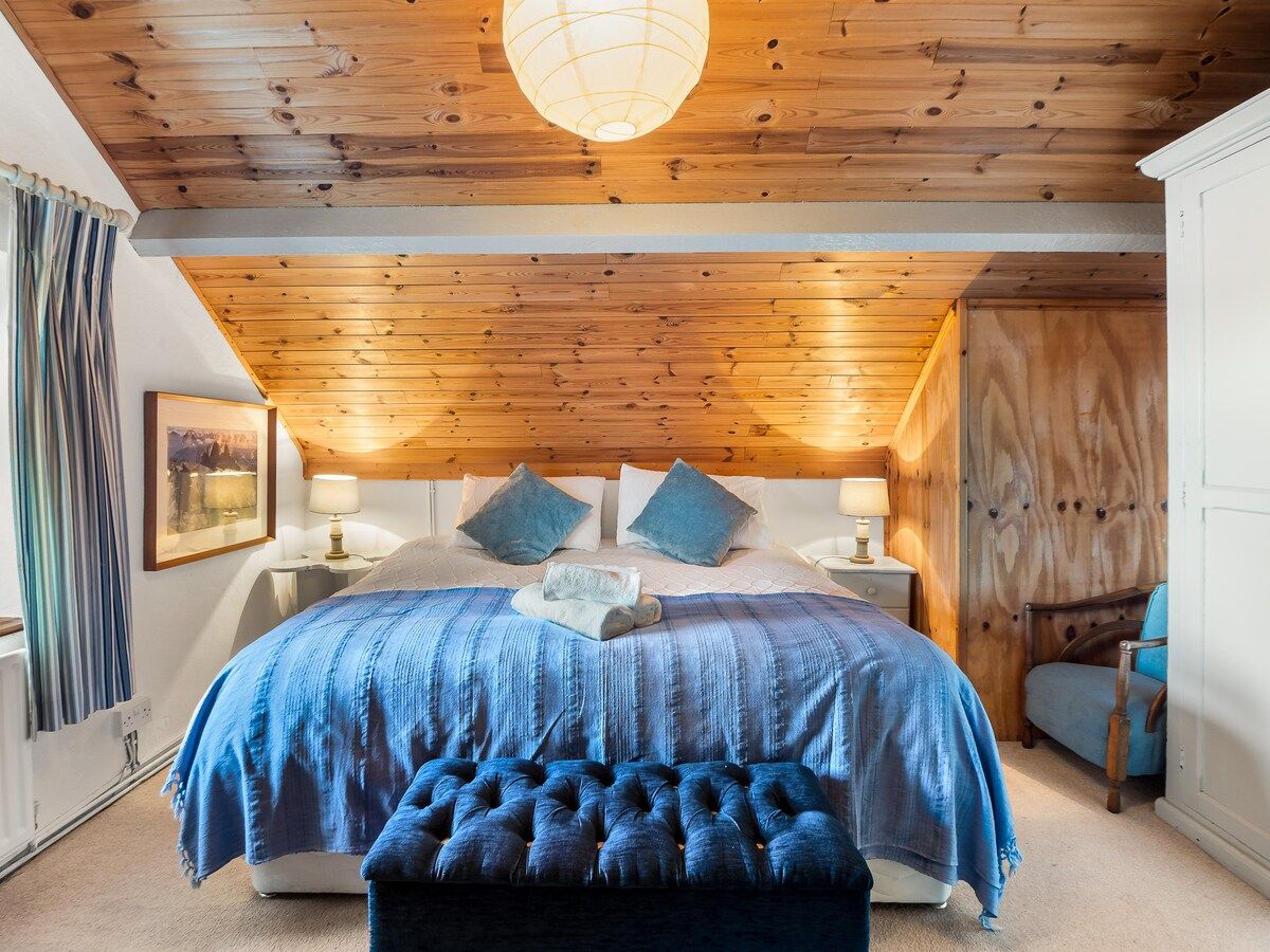A bedroom with a wooden ceiling, a bed with a blue bedspread, two bedside tables with lamps, and a chair.