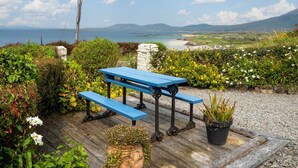 Une terrasse en bois avec une table de pique-nique bleue et des bancs, offrant une vue sur un paysage côtier avec des montagnes à l’horizon.